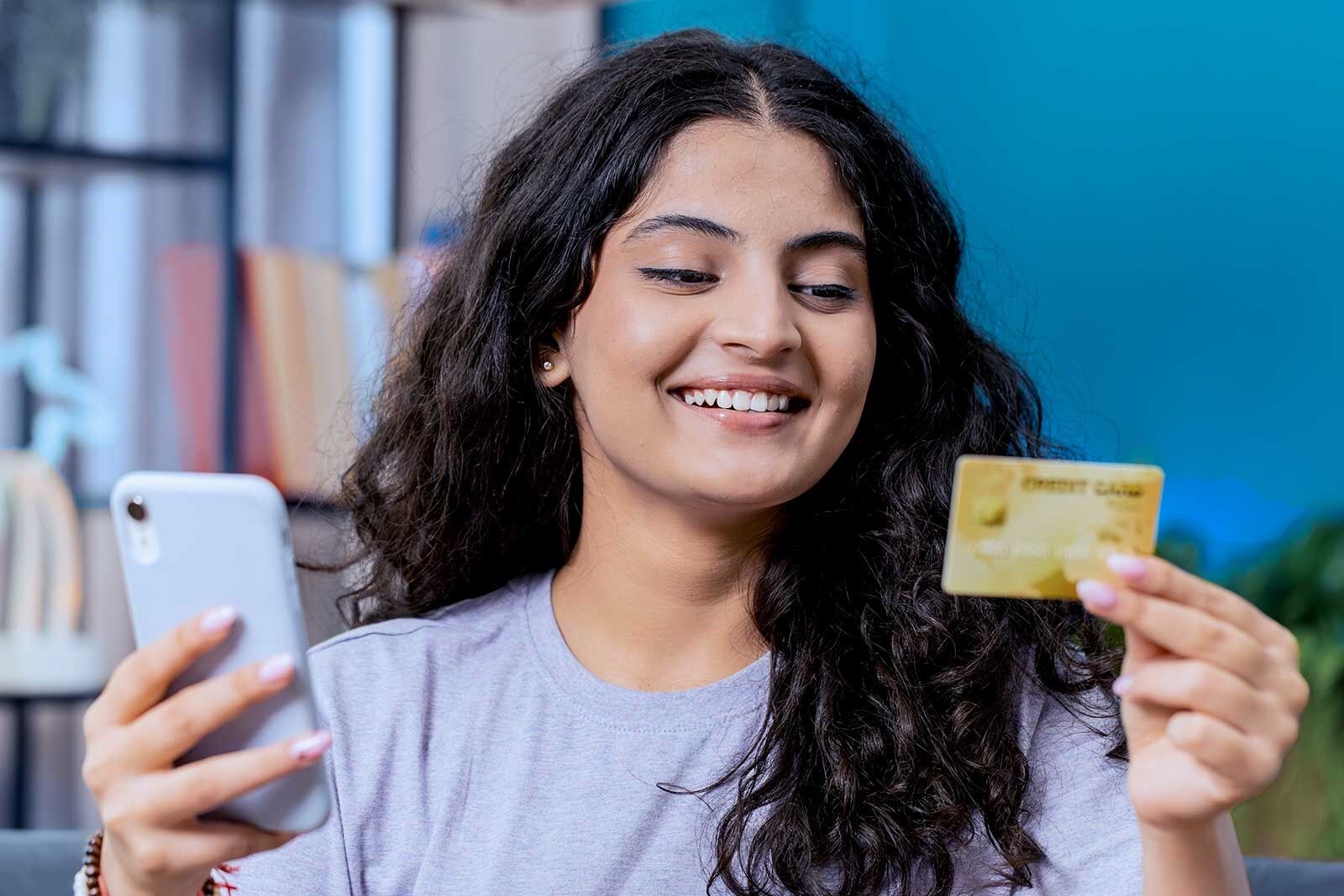 Uma jovem de cabelos escuros e cacheados sorri enquanto segura um cartão de crédito e um telefone celular.