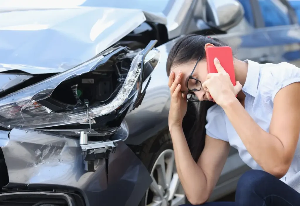 mulher ao lado da frente do seu carro batido com a mão na testa enquanto liga para o socorro