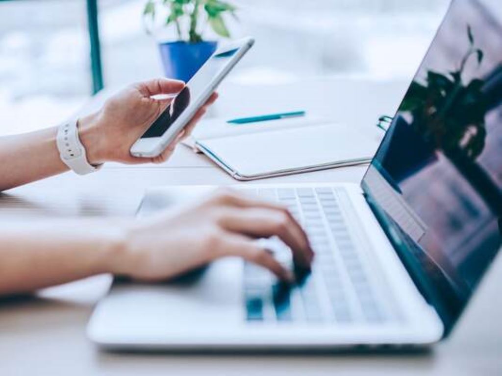 Pessoa mexendo no celular com a mão esquerda e no computador com a mão direita em cima da mesa, com bloco de notas e planta ao fundo
