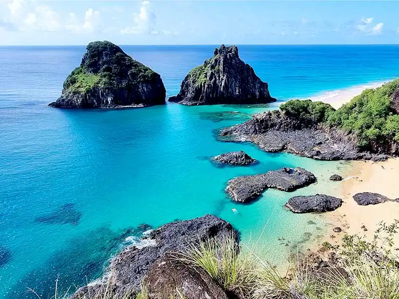 Vista aérea de praia paradisíaca com águas cristalinas turquesa, rochas escuras e duas ilhas rochosas no oceano.