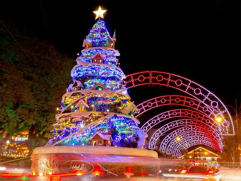 Uma grande árvore de Natal iluminada, coberta de neve artificial e pequenas casas, está localizada em uma praça, ladeada por arcos de luzes coloridas. O cenário noturno é vibrante e festivo.