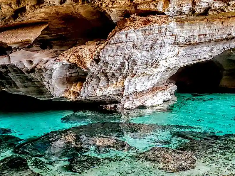 Água turquesa brilhante em uma caverna de pedra calcária, com paredes de rocha marrom e branca e uma abertura escura na formação rochosa.