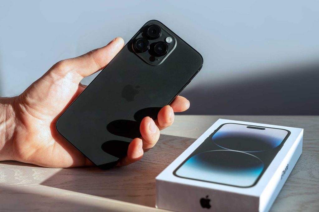 Uma mão segurando um iPhone 12 Pro Max preto, com sua caixa na mesa ao lado.