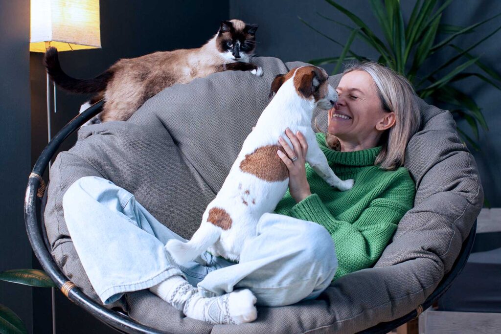 Mulher sorrindo com cachorro e gato em um sofá confortável.