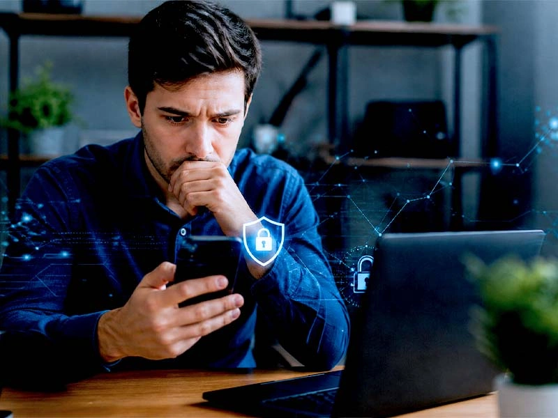 Homem preocupado olha celular ao lado de notebook com ícones brilhantes de cadeado e escudo de proteção.