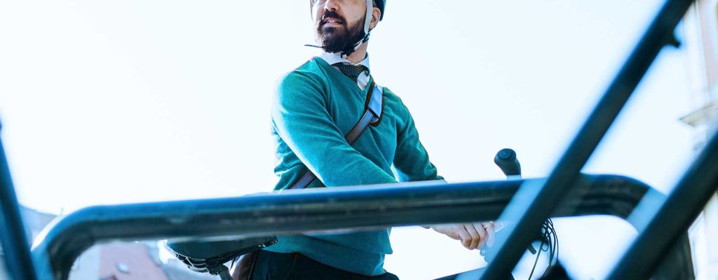 Homem barbudo com capacete de bicicleta azul, vestindo um suéter verde, está parado perto de sua bicicleta em um suporte de bicicleta ao ar livre.