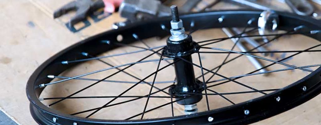 Roda de bicicleta preta com raios e cubo, sobre uma superfície com várias ferramentas espalhadas ao fundo.