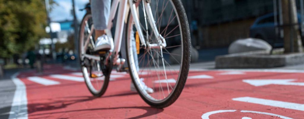 Uma pessoa pedala uma bicicleta branca em uma ciclovia vermelha com marcações de bicicletas pintadas no chão.