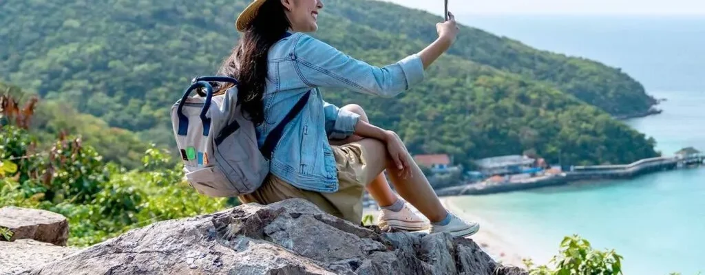 Uma mulher sorridente com mochila tira uma selfie em um penhasco com vista para uma praia tropical.