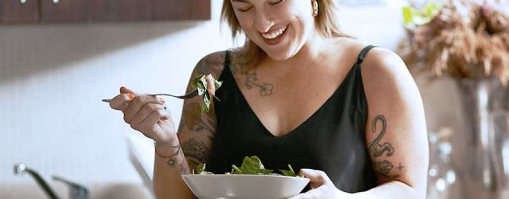 Conteudo_Orientação-nutricional-o-que-é-como-fazer-benefícios Uma mulher sorridente com tatuagens nos braços está sentada em sua cozinha, comendo uma salada de uma tigela.