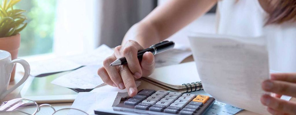Uma mulher revisando documentos e usando uma calculadora para fazer cálculos financeiros em sua mesa. Há uma caneta, óculos e uma planta em um vaso ao lado dela.