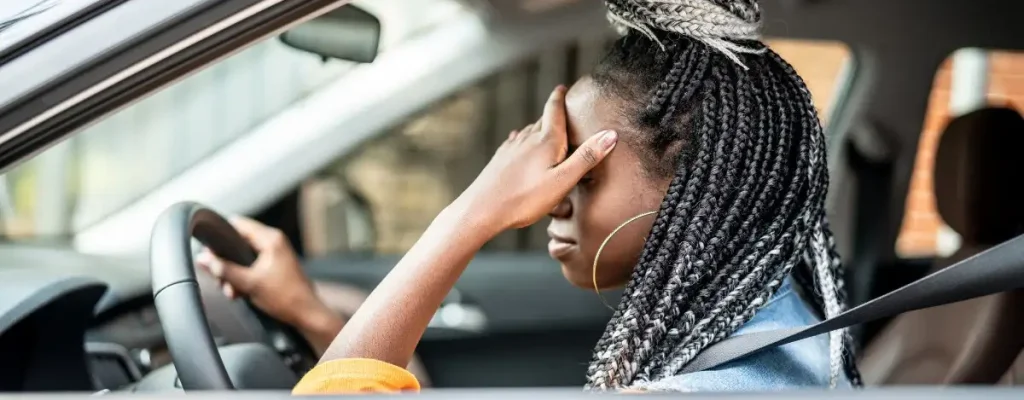 moça sentada no carro no banco de motoristas, com as mãos na testa e uma expressão frustrada
