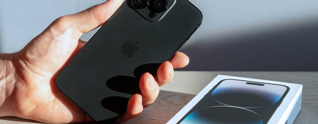 Uma mão segurando um iPhone 12 Pro Max preto, com sua caixa na mesa ao lado.